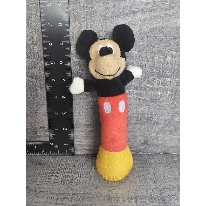 May include: A plush Mickey Mouse toy with black ears, a black torso, red legs, and a yellow bottom. The toy has white gloves and two white oval buttons on the red portion. The toy is approximately 20 cm tall.