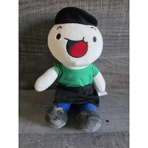 May include: A plush doll with a white head, black beret, and a green shirt. The doll has a large red mouth and black eyes. It is wearing a black skirt and blue pants. The doll's feet are gray.