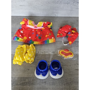 May include: A clown costume for a doll, featuring a red and white polka dot dress with a rainbow trim, yellow and white polka dot pants, blue shoes, a red and white polka dot hat, and a red and yellow clown mask.