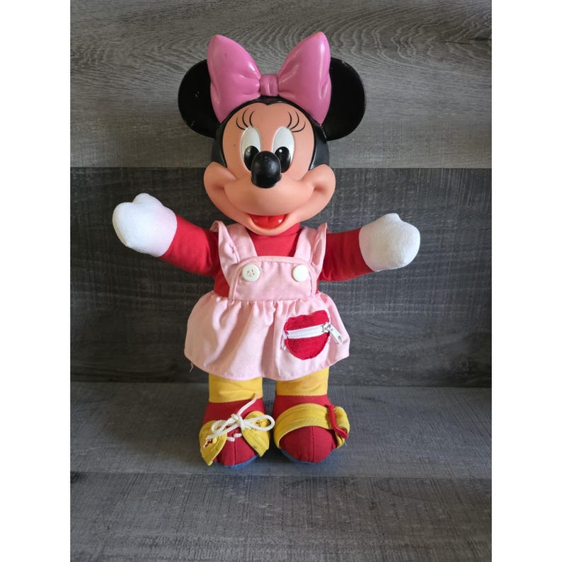 May include: A vintage Minnie Mouse plush doll. The doll has a pink bow, black ears, and a red top. She wears a pink dress with white buttons and a red pocket. Her shoes are red and yellow.