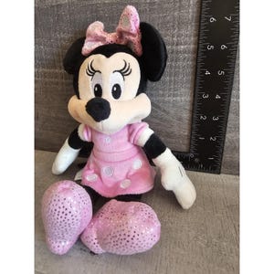 May include: A pink and black plush toy of Mickey Mouse's girlfriend Minnie Mouse. She is wearing a pink dress with white polka dots and a pink bow with silver glitter.