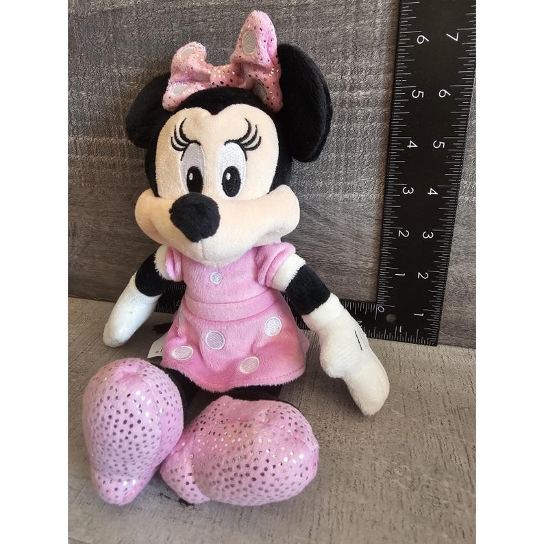 May include: A pink and black plush toy of Mickey Mouse's girlfriend Minnie Mouse. She is wearing a pink dress with white polka dots and a pink bow with silver glitter.
