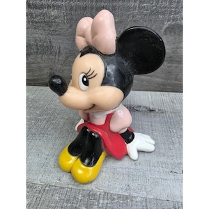 May include: A vintage Minnie Mouse figurine. The figure is in a seated position, wearing a red dress, pink bow, and yellow shoes. The figurine has black ears and a black nose. The figure is made of a glossy material.