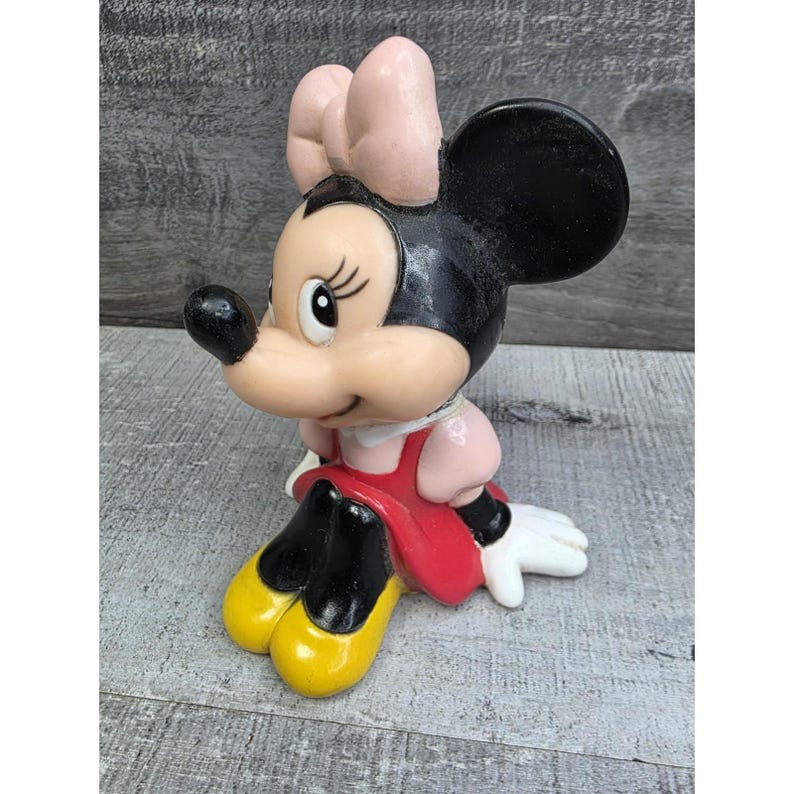 May include: A vintage Minnie Mouse figurine. The figure is in a seated position, wearing a red dress, pink bow, and yellow shoes. The figurine has black ears and a black nose. The figure is made of a glossy material.