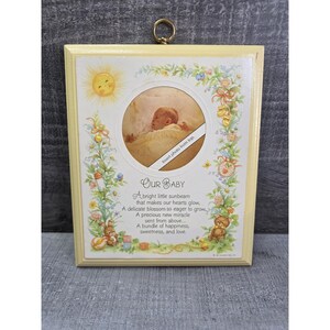May include: A framed baby picture with a yellow border. The picture has a circular opening for a photo, surrounded by floral and sun illustrations. Text reads "Our Baby" and a poem about a newborn.