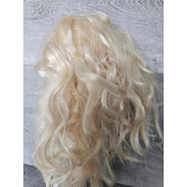 以下が含まれることがあります： 明るいブロンドの波状のドールヘアの塊。髪は淡いクリーム色で、柔らかく質感のある外観です。カールはゆるく流れており、ボリューム感のある外観を作り出しています。髪は合成繊維のようです。