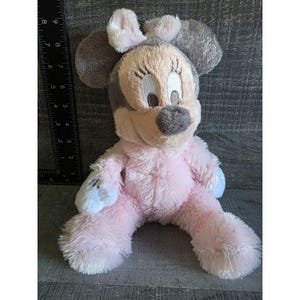 May include: A plush Minnie Mouse doll in a seated position. The doll features a pink body, gray ears, and a light pink bow. The face is beige with black eyelashes and a gray nose. The doll is approximately 8 inches tall.