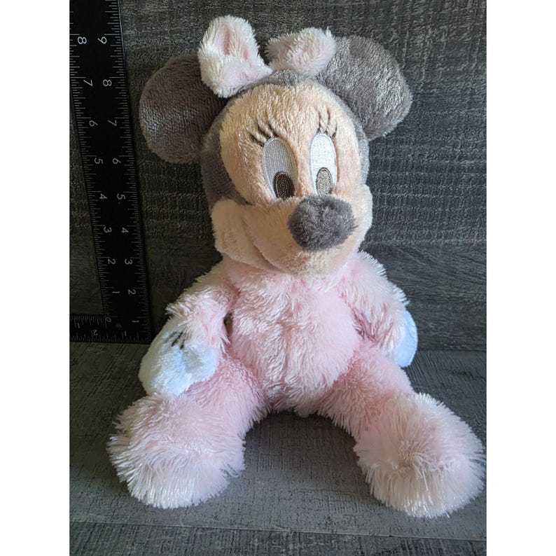 May include: A plush Minnie Mouse doll in a seated position. The doll features a pink body, gray ears, and a light pink bow. The face is beige with black eyelashes and a gray nose. The doll is approximately 8 inches tall.
