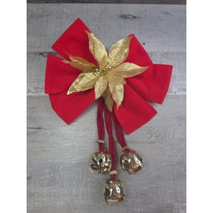 Puede incluir: Un lazo navideño festivo con un gran lazo de terciopelo rojo, una flor de pascua dorada y tres campanas doradas colgando de cordones rojos. Una decoración navideña clásica.