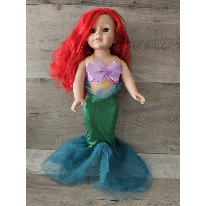 Puede incluir: Una mu&ntilde;eca de pelo rojo con un disfraz de sirena verde y morado. El disfraz incluye un top morado con un lazo y una cola verde con una falda de tul azul.