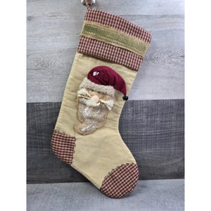 May include: Tan Christmas stocking with a Santa Claus face applique. The stocking has a burgundy and tan plaid pattern on the cuff and toe, with a green burlap ribbon accent. The Santa face is made of burlap and has a burgundy hat.