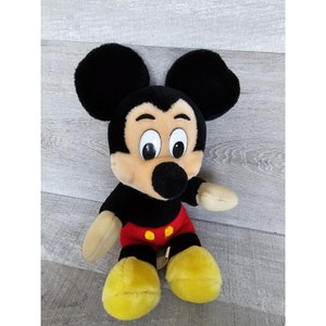 May include: A plush Mickey Mouse toy with black ears, a black nose, and large, white eyes. The toy is wearing red shorts with two yellow buttons.