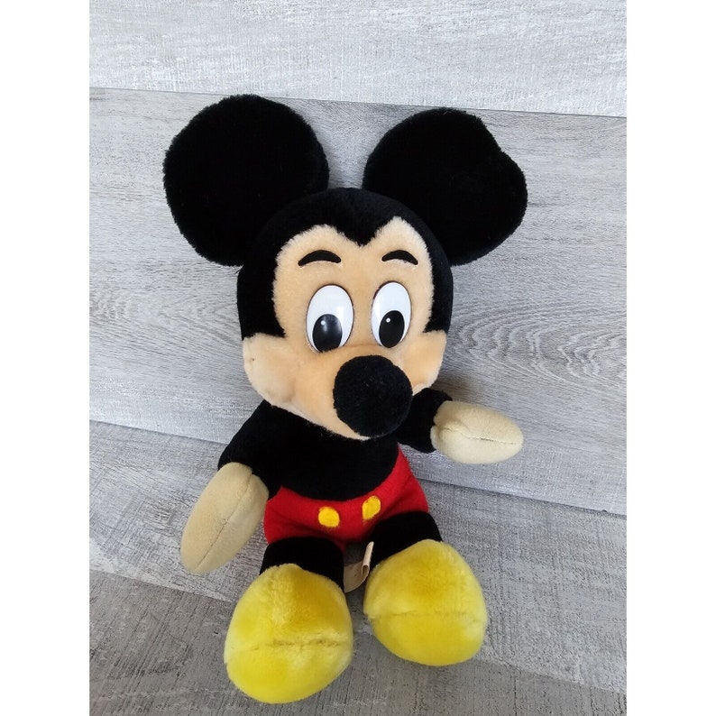 May include: A plush Mickey Mouse toy with black ears, a black nose, and large, white eyes. The toy is wearing red shorts with two yellow buttons.