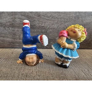 Peut inclure: Deux poupées Cabbage Patch Kids, l'une debout et tenant un cornet de glace, l'autre faisant un poirier. La poupée debout porte une robe bleue avec des bordures blanches et a les cheveux blonds. La poupée faisant un poirier porte un pantalon bleu et un haut rouge et blanc.