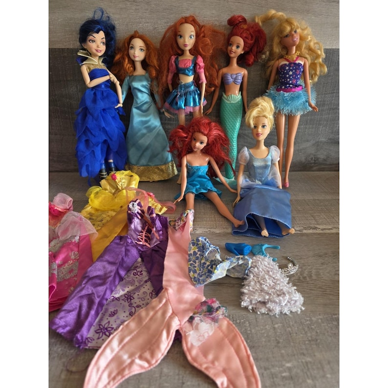Op de afbeelding: Een verzameling modepoppen met verschillende kapsels en outfits. De poppen dragen kleurrijke jurken, rokken en tops. Extra poppenkleding en accessoires zijn ook inbegrepen, zoals een zeemeerminstaart en een zilveren tiara.