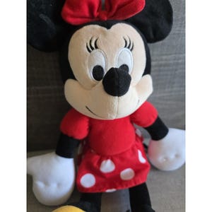 May include: A plush Minnie Mouse doll with a red dress with white polka dots, a red bow, and black ears. The doll has white gloves and a black nose. The doll is a popular character from Disney.