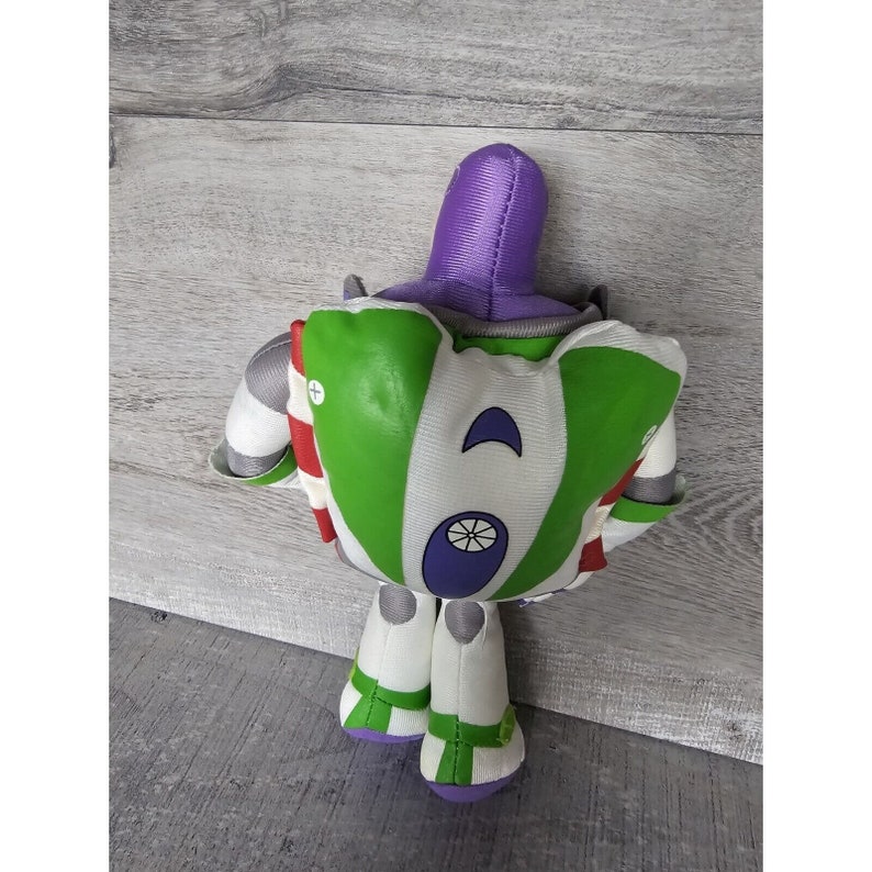 Peut inclure: Un jouet en peluche de Buzz l'&Eacute;clair de la franchise Toy Story. Le jouet est vert, blanc et violet avec une bande rouge. Le jouet est assis sur une surface en bois.