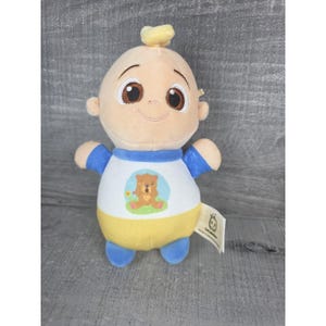 May include: A plush doll of a cartoon baby with a smiling face, brown eyes, and a tuft of yellow hair. The doll wears a white shirt with a bear graphic, blue sleeves, and a yellow bottom. A tag is attached.