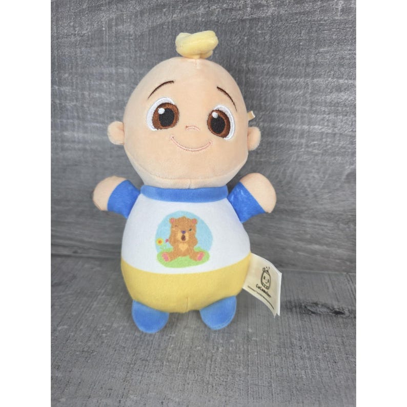 May include: A plush doll of a cartoon baby with a smiling face, brown eyes, and a tuft of yellow hair. The doll wears a white shirt with a bear graphic, blue sleeves, and a yellow bottom. A tag is attached.