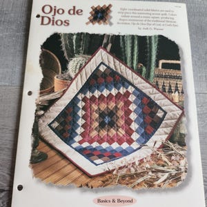 May include: A printed instructional pattern sheet titled Ojo de Dios by Jodi G. Warner. The cover features a square quilted wall hanging with a geometric pattern in shades of blue, red, brown, and cream, with the text Basics & Beyond at the bottom.