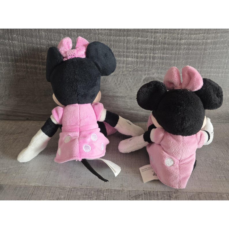 May include: Two Minnie Mouse plush dolls. The dolls have black ears and heads, pink bows, and pink dresses with white polka dots. They have white gloves and black and white arms. The dolls are on a gray surface.