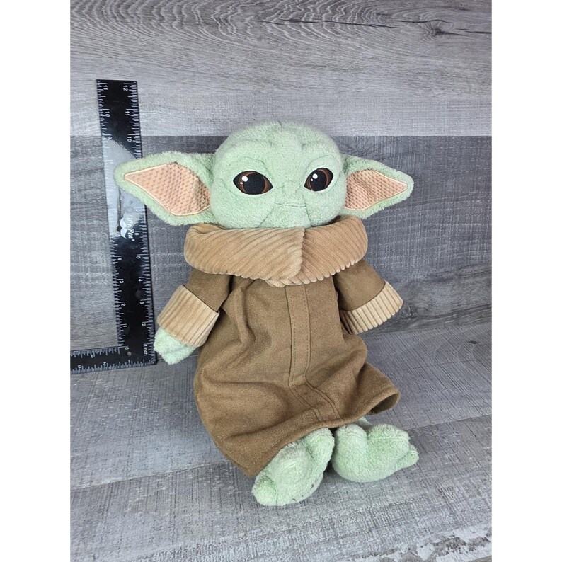 May include: A plush doll of a character with large ears, big eyes, and a tan and brown outfit. The doll is approximately 12 inches tall. The doll has a light green head and feet, with tan ears and a brown coat. The doll is sitting on a gray wooden surface.