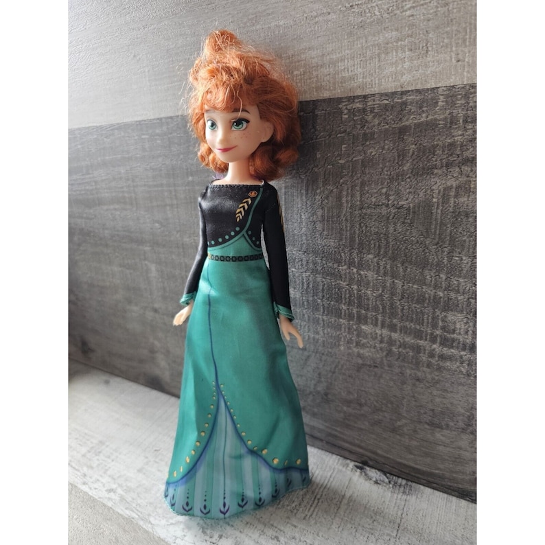 May include: A red-haired doll wearing a teal and black dress with gold accents. The doll has a long, flowing dress with a black bodice and a teal skirt. The dress is decorated with gold accents, including a leaf design on the bodice and a pattern of dots and lines on the skirt.