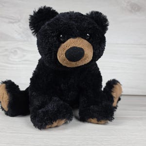 May include: A plush black bear toy with soft, textured fur, featuring a tan-coloured muzzle, a round black nose, and matching tan accents on the soles of its feet. The bear has dark, round eyes and is depicted in a seated position with its limbs extended.