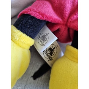 Puede incluir: Un juguete de peluche de Mickey Mouse con pantalones cortos rojos, brazos y piernas negros y guantes amarillos. El juguete tiene una etiqueta blanca con la palabra "Disney" impresa en ella.