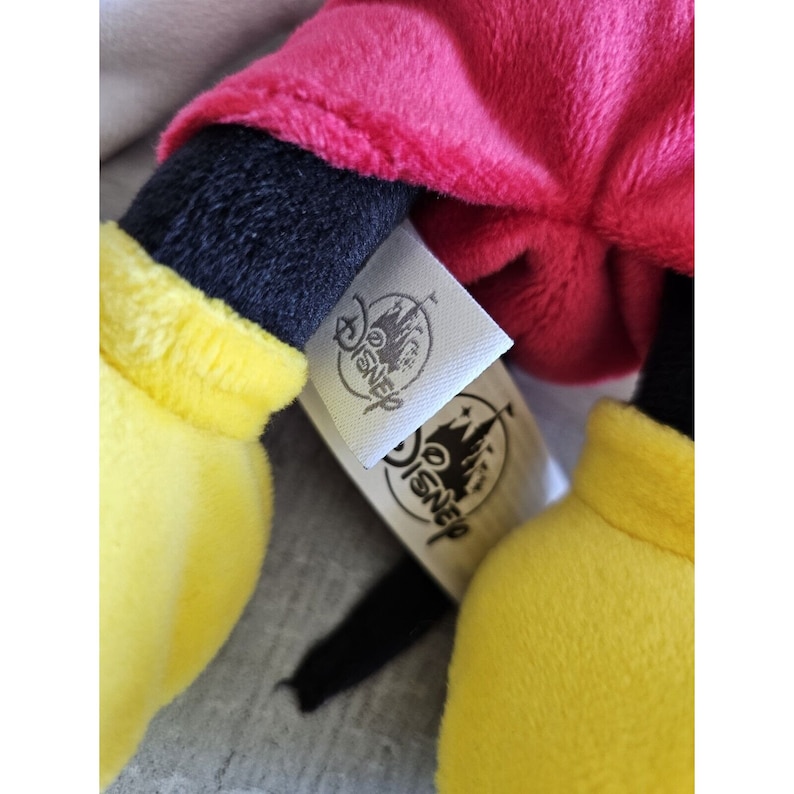Puede incluir: Un juguete de peluche de Mickey Mouse con pantalones cortos rojos, brazos y piernas negros y guantes amarillos. El juguete tiene una etiqueta blanca con la palabra "Disney" impresa en ella.