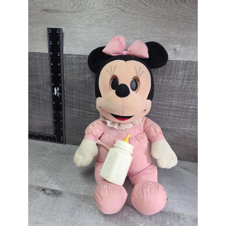 Puede incluir: Mu&ntilde;eca de peluche de Minnie Mouse con un vestido y lazo rosa, sosteniendo un biber&oacute;n blanco. La mu&ntilde;eca tiene orejas negras, cara color crema y guantes blancos. La mu&ntilde;eca mide aproximadamente 30 cm de alto.