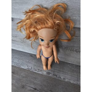 May include: A doll with red hair, blue eyes and a blue swimsuit. The doll is sitting on a wooden surface.