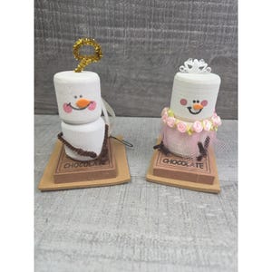 May include: Two marshmallow snowman ornaments. One has a gold tinsel top and the other has a tiara and pink tulle skirt. Both have a smiling face and sit on a chocolate bar base with the word "CHOCOLATE".