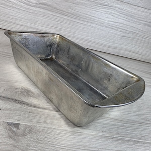Peut inclure: Un moule à pain rectangulaire en métal avec une finition argentée patinée. L'ustensile de cuisson présente un intérieur profond et ouvert avec des parois légèrement inclinées et une base plate, montrant des traces d'usure et de métal.