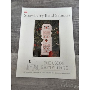 May include: A cross-stitch pattern for a sampler featuring a strawberry band with the alphabet, numbers, and a heart. The pattern is titled "Strawberry Band Sampler" and is from Hillside Samplings.