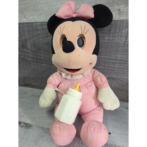 Puede incluir: Mu&ntilde;eca de peluche vintage de Minnie Mouse con un vestido rosa y un lazo a juego. La mu&ntilde;eca tiene orejas negras, un cuello blanco y sostiene un biber&oacute;n blanco con una tetina amarilla. La mu&ntilde;eca mide aproximadamente 30 cm de altura.