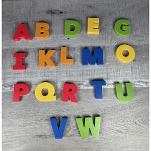 May include: Colorful magnetic alphabet letters in various colors, including red, orange, yellow, green, and blue. The letters are arranged on a gray wooden surface. The letters are capital letters.