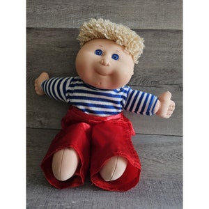 以下が含まれることがあります： 金髪で青い目をした、笑顔のキャベツパッチキッズ人形。人形は青と白のストライプのシャツと赤いズボンを着ています。