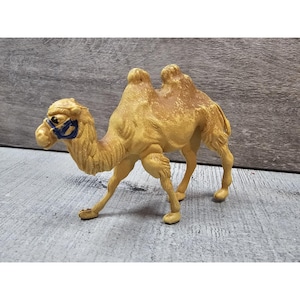 Op de afbeelding: Een bruin plastic kameelbeeldje met twee bulten en een blauwe teugel. De kameel is afgebeeld in een loopstand, met uitgestrekte poten. Het beeldje heeft een getextureerde vacht en staat tegen een grijze achtergrond.