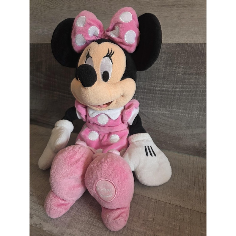 May include: A Minnie Mouse soft toy, dressed in a pink dress with white polka dots and a matching bow. The toy has black ears, a black nose, and white gloves. The toy is sitting on a wooden surface.