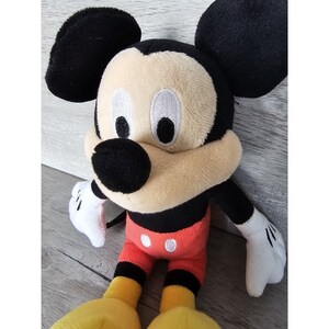Puede incluir: Un juguete de peluche de Mickey Mouse con orejas negras, nariz negra y ojos blancos. El juguete lleva pantalones cortos rojos con dos botones blancos y zapatos amarillos.