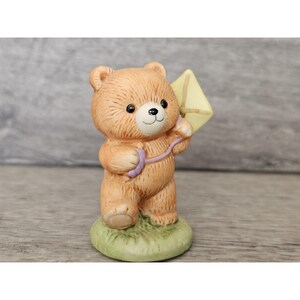 Puede incluir: Una figurita de cerámica de un oso de peluche marrón que sostiene una cometa amarilla. El oso está de pie sobre una base verde.