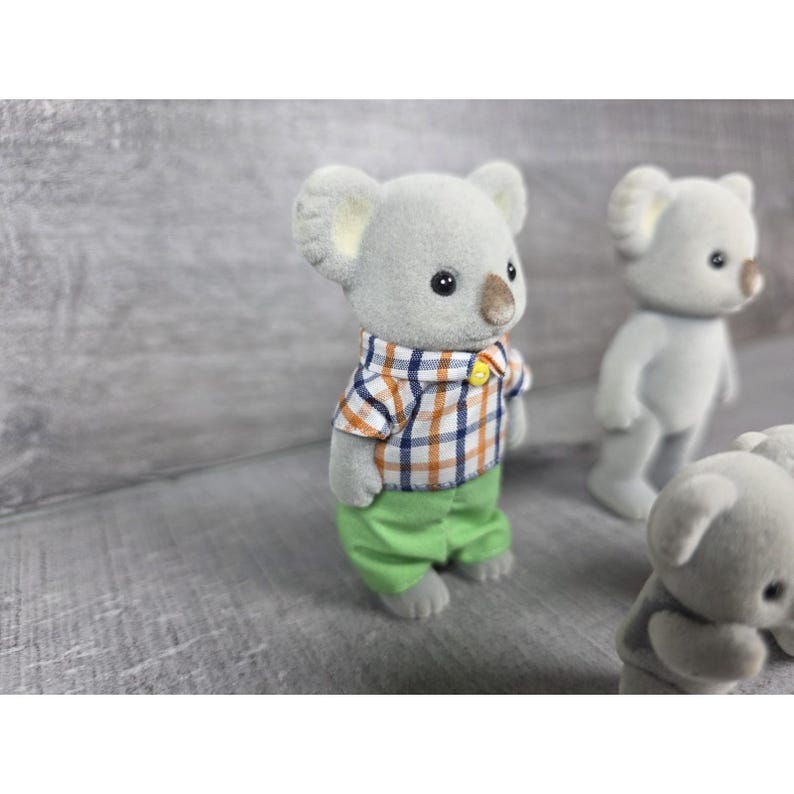 May include: A collection of miniature koala bear figurines. One koala wears a plaid shirt with orange, blue, and white squares, and green pants. Other koalas are visible in the background, with gray fur and small black eyes. The figurines are on a gray surface.