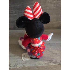May include: A Minnie Mouse plush toy, viewed from the back, features a black head and ears, a red and white striped bow, and a red dress with a colorful geometric pattern. The toy has white gloves and is seated.