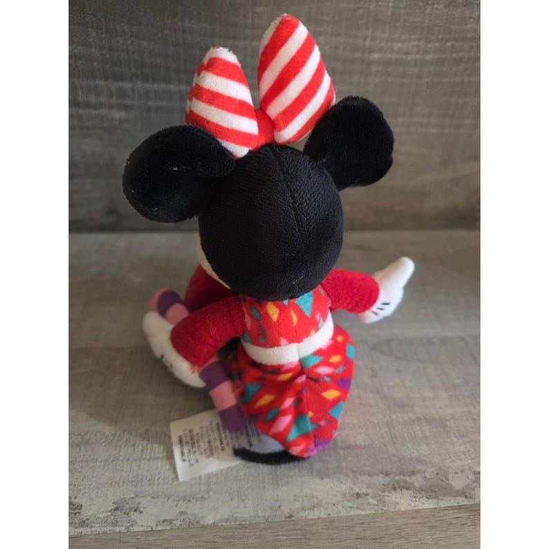 May include: A Minnie Mouse plush toy, viewed from the back, features a black head and ears, a red and white striped bow, and a red dress with a colorful geometric pattern. The toy has white gloves and is seated.