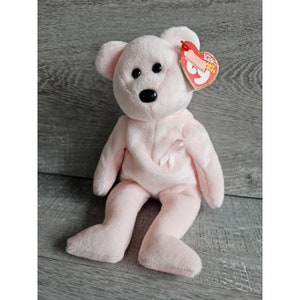 Könnte beinhalten: Rosafarbener Beanie Baby Teddybär mit einem rosa Band auf der Brust. Der Bär hat schwarze Augen und eine schwarze Nase. Der Bär sitzt auf einer Holzoberfläche. Das Etikett lautet "Ty Inc. Beanie Babies"