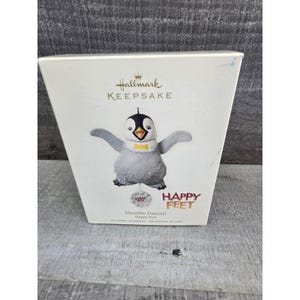 Könnte beinhalten: Hallmark Keepsake Weihnachtsschmuck mit einem Plüschpinguin mit ausgestreckten Armen und einer gelben Fliege. Der Schmuck befindet sich in einer weißen Schachtel mit den Aufschriften "Happy Feet" und "Mumble Dances!"
