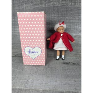 May include: A vintage Effanbee doll in a red coat and white dress, with a matching hat. The doll is standing next to a pink box covered in white hearts, with the Effanbee logo. The doll's outfit includes black shoes.