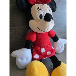 May include: A plush Minnie Mouse doll. The doll has a cream-colored face, black ears, and a red bow. She wears a red dress with white polka dots, black arms and legs, and yellow shoes. The doll's hands are white.