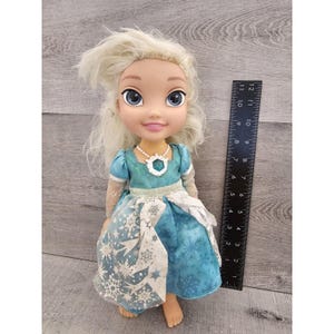 May include: A blonde doll wearing a blue dress with a snowflake pattern. The doll has blue eyes and is wearing a necklace with a blue gem. The doll is about 11 inches tall.